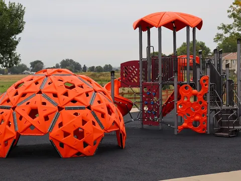 The new playground on the north side of Sunrise Park: an orange climbing dome and a slide tower with an orange canopy on a black rubber surface, with a rail fence and farm fields behind